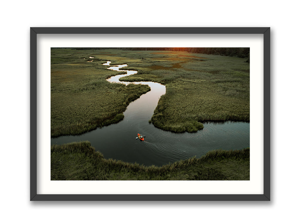 A framed photograph depicting a kayak on a winding waterway amidst green marshlands, with a pink sunset in the background.