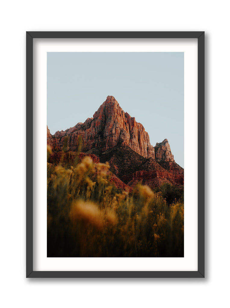 A framed print depicting a landscape photograph of Zion National Park, featuring a prominent sandstone spire known as the Watchman, with autumn-colored vegetation in the foreground.