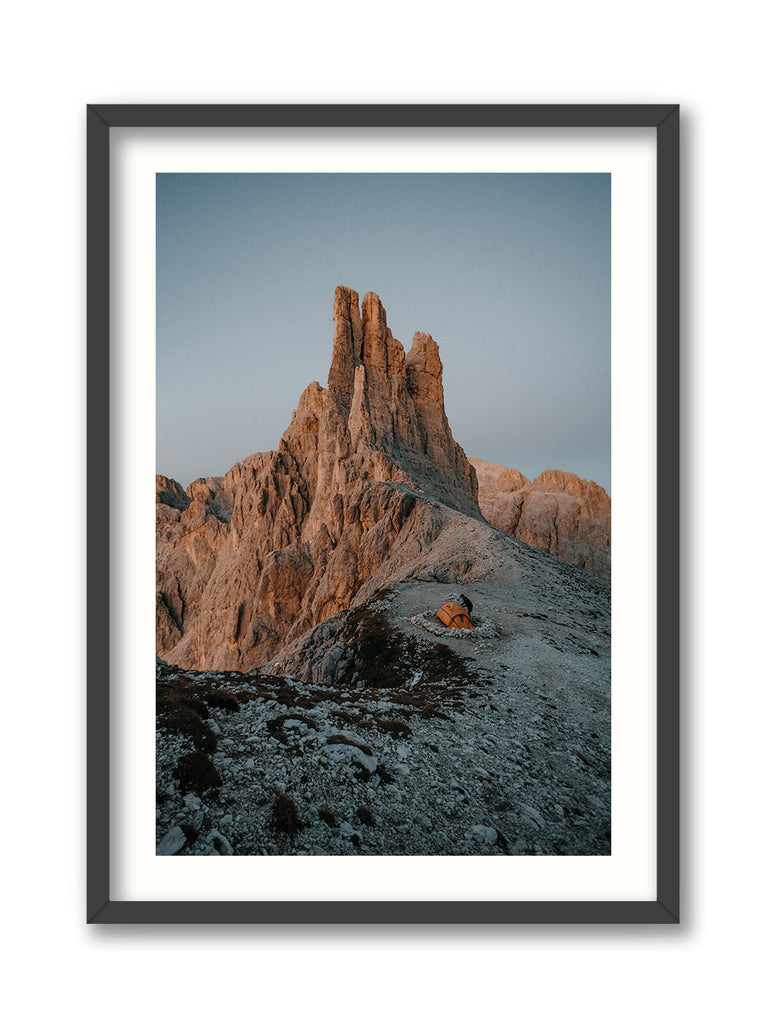 A framed print depicting the Vajolet Towers mountain range in the Dolomites, with a hiker setting up a tent in the foreground.