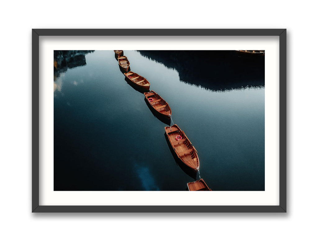 A framed artwork featuring a photograph of wooden boats on water with a backdrop of lush greenery and mountains.