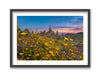 A framed artwork depicting a landscape with yellow wildflowers in the foreground and tall rock formations, known as the Trona Pinnacles, in the background against a pink and purple sky during sunset.